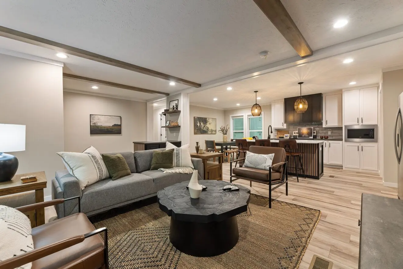 Open-plan living room and kitchen of a finished Lucky Ranch Trail model home — exposed wood ceiling beams, gray sectional, sculptural round coffee table on a jute rug, leather accent chair, pendant-lit kitchen island, and white cabinetry